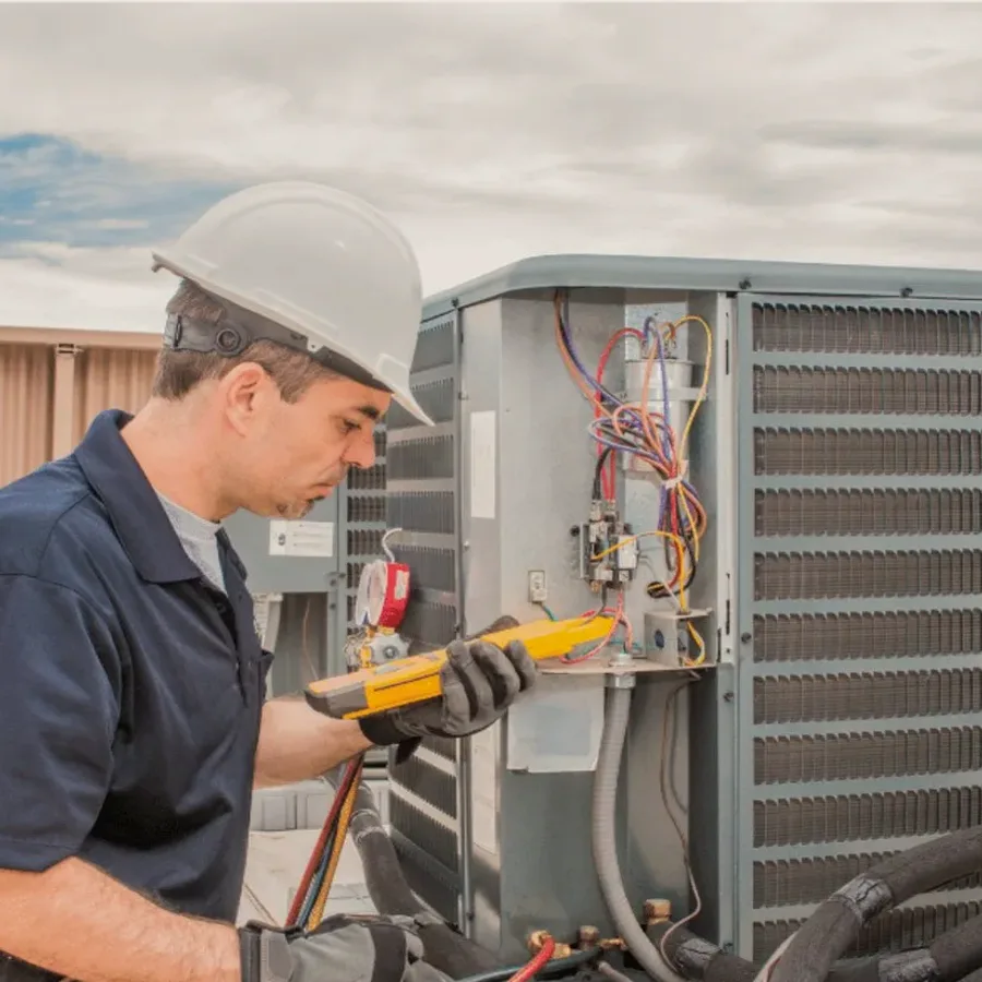 HVAC technician in a hard hat testing an outdoor air conditioner with a multimeter